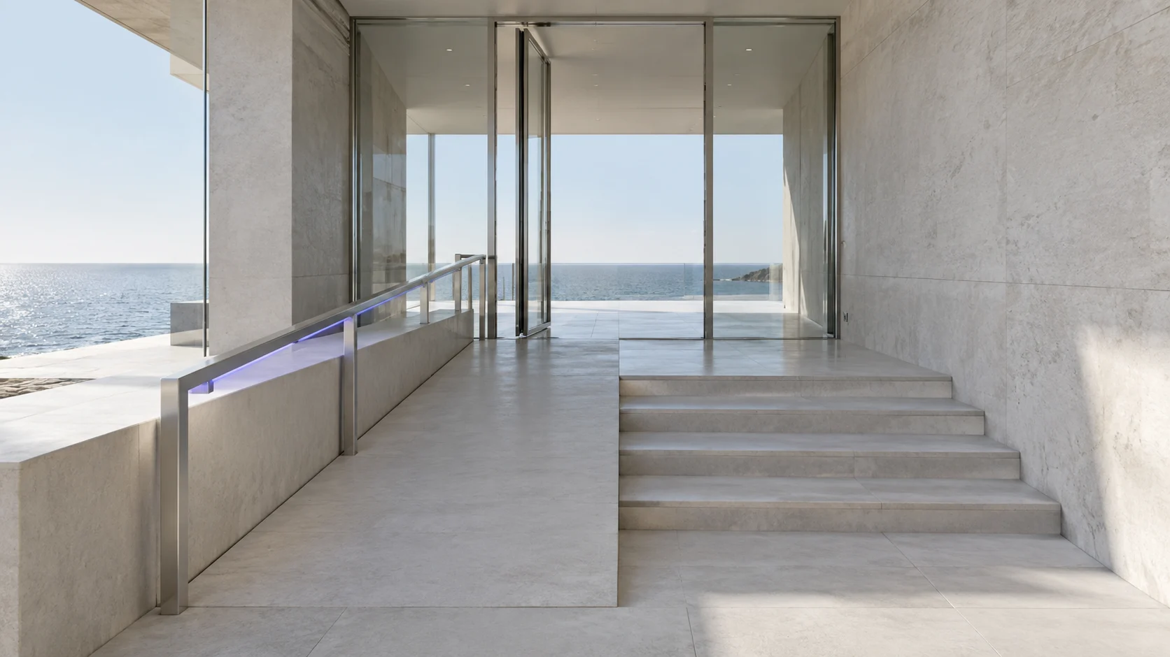 Arriving at a glazed entrance where a shallow ramp and a short flight of steps reach the same open door, in concrete with a brushed steel handrail lit violet-blue underneath, and the sea beyond the glass.