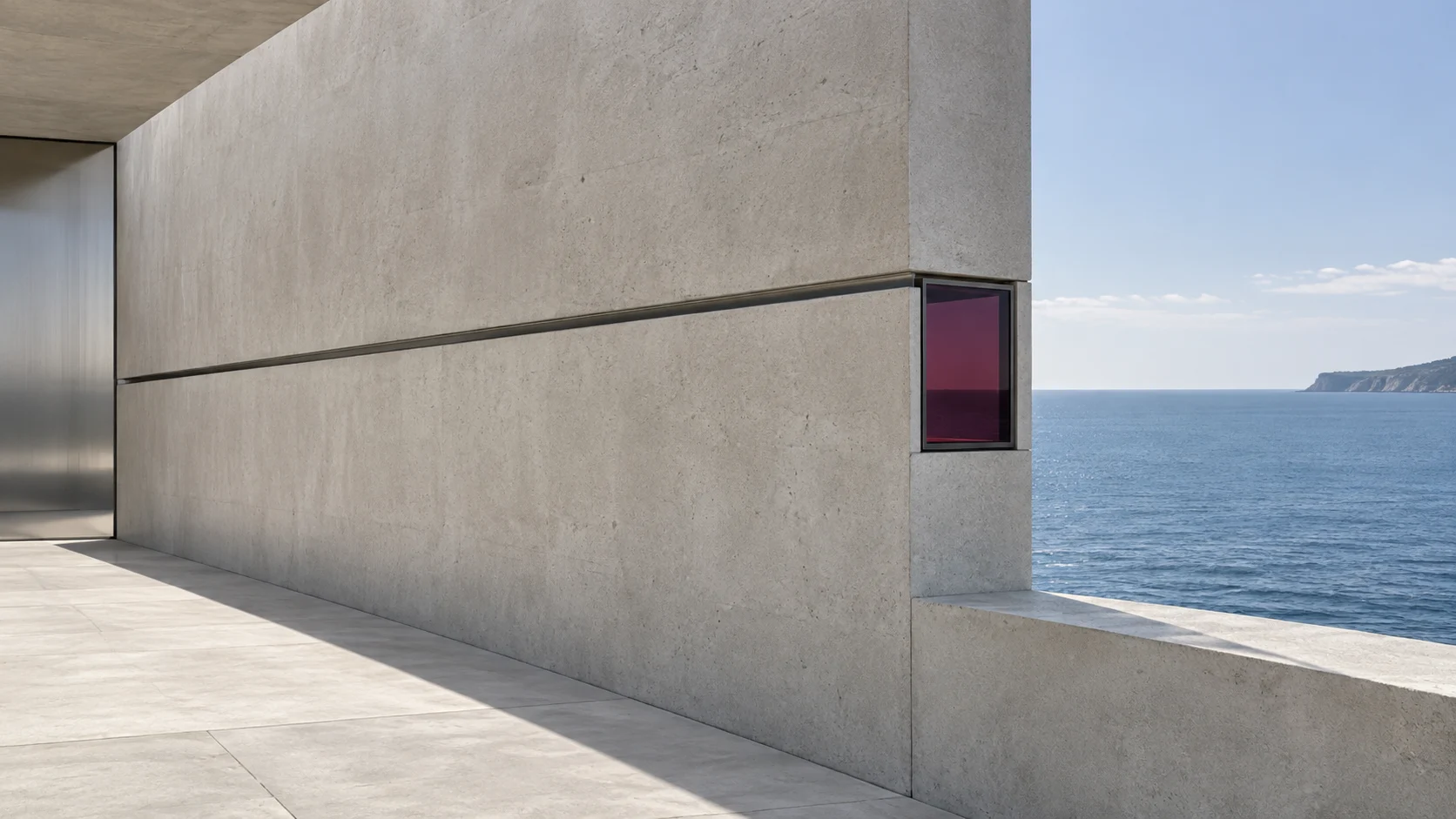 A long concrete wall with one horizontal steel line running to a small window of deep red glass at its end, and the sea beyond.