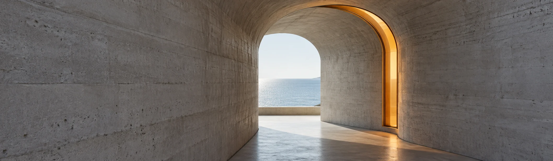 A board-marked concrete passage ending in an arched opening onto the sea, the inside of the arch lined with amber light.