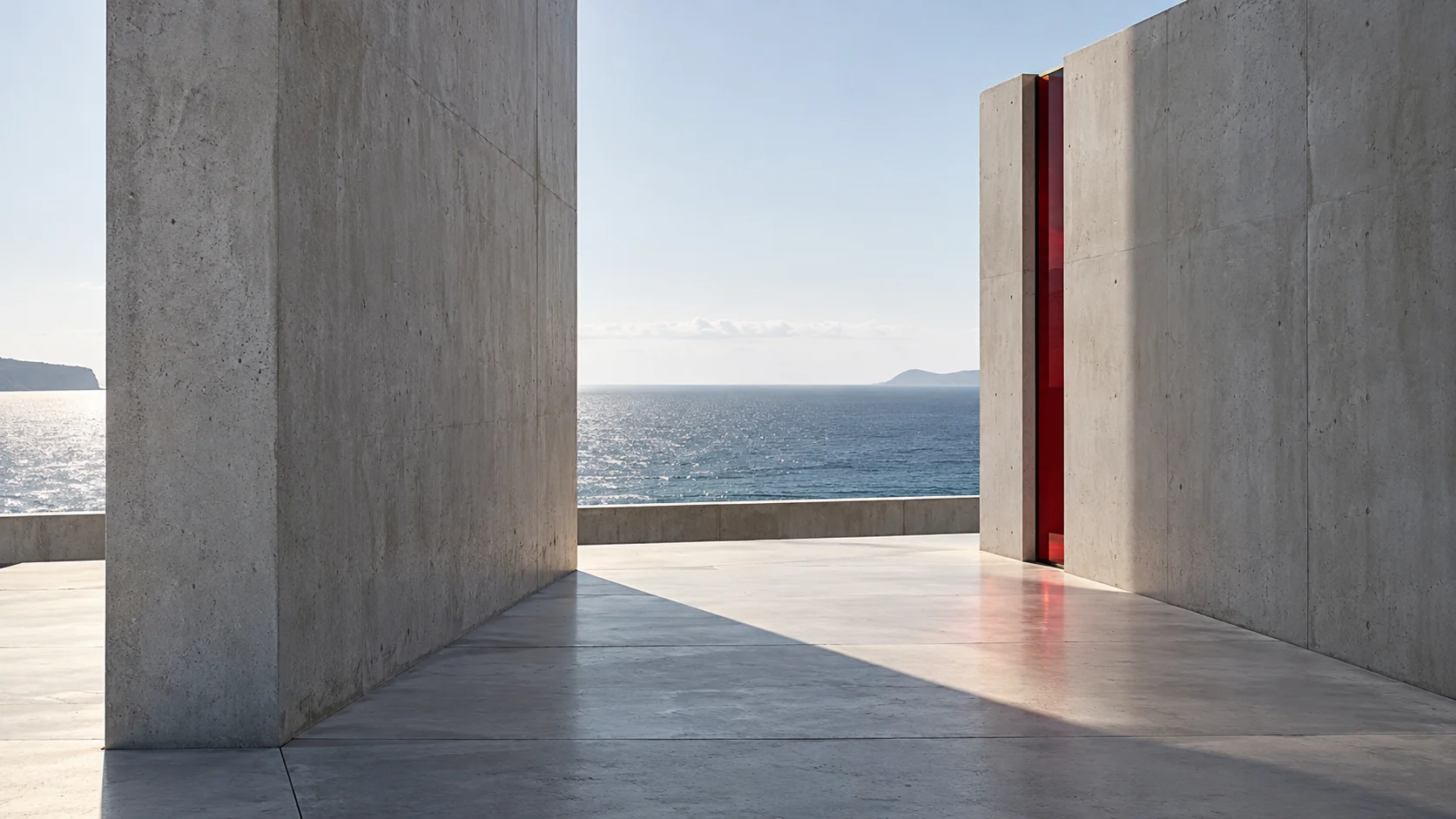 Two concrete walls with an open gap between them onto the sea. The near wall’s shadow lies across the floor and stops short of the far wall, which has a strip of ruby glass at its edge.