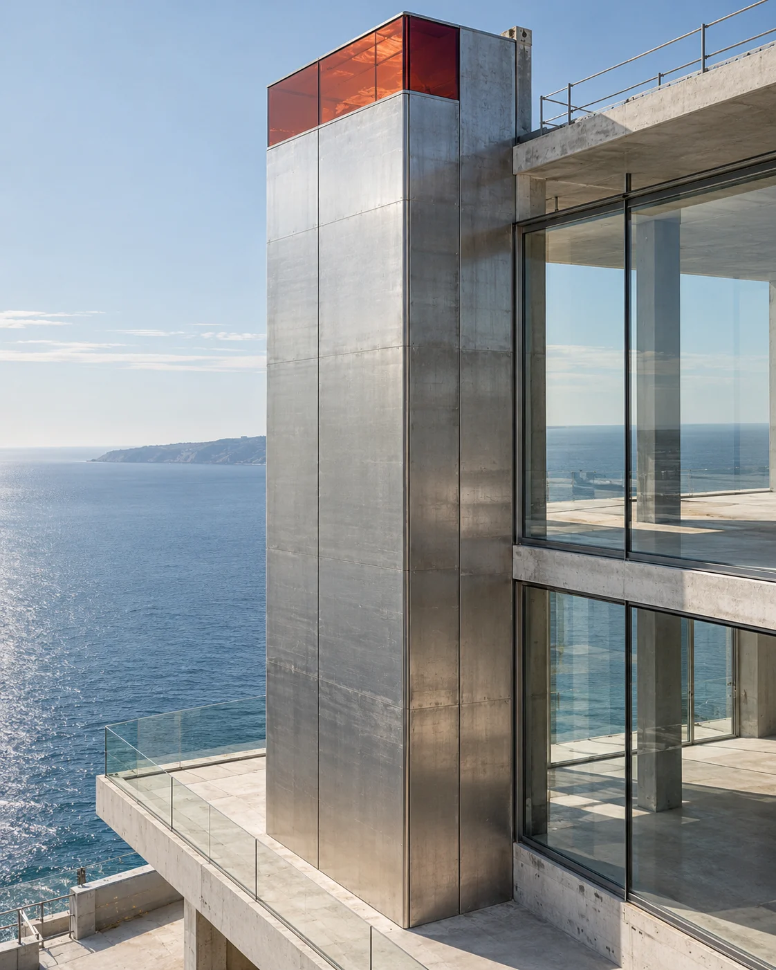 A tall steel-clad core standing clear beside the glazed floors of the building above the sea, a band of carnelian glass at its top the only colour.