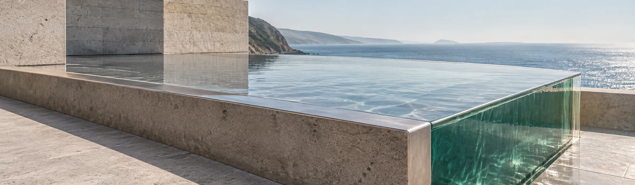 The corner of the glass-sided pool as a band: the water level with its stone and steel rim, the emerald glass corner at the right, the sea and a headland beyond.