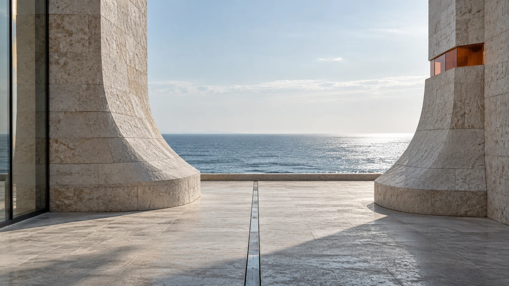 A stone court open to the sea: two flared travertine piers stand on either side of one inlaid steel line, and a carnelian glass band is set into the pier at the right.