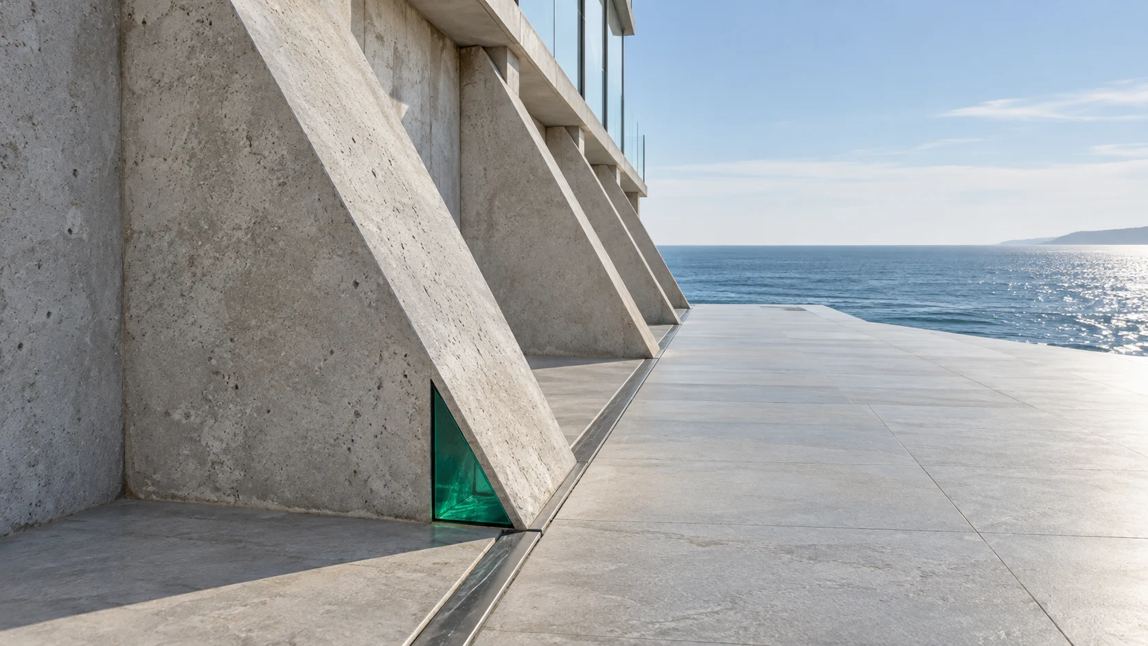A row of concrete buttresses standing along one floor joint beside a terrace above the sea, with an emerald glass wedge set at the foot of the nearest.