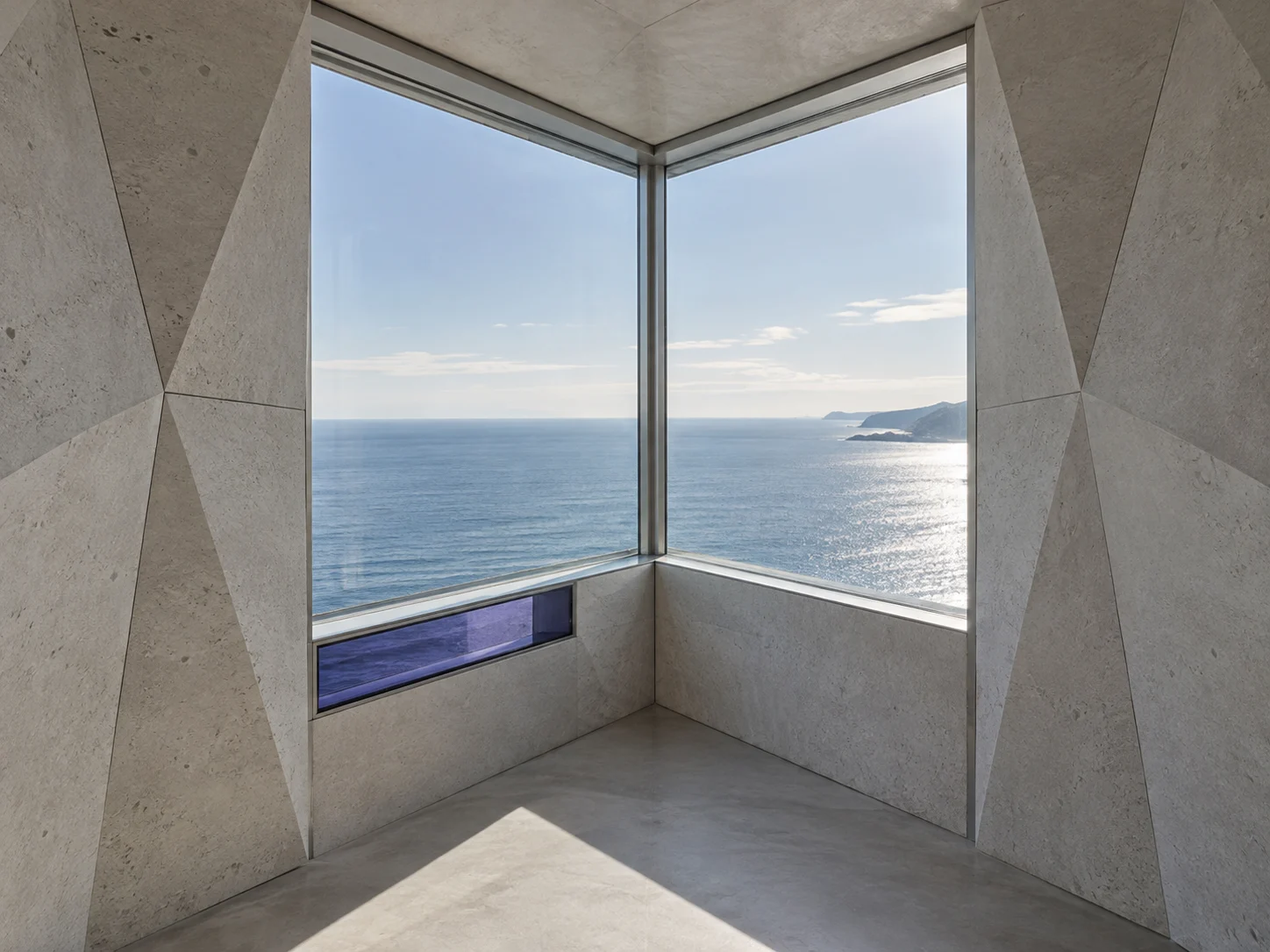 A glazed corner in a room of faceted stone panels, the sea beyond the glass, with one violet-blue glass panel set below the window at the left.